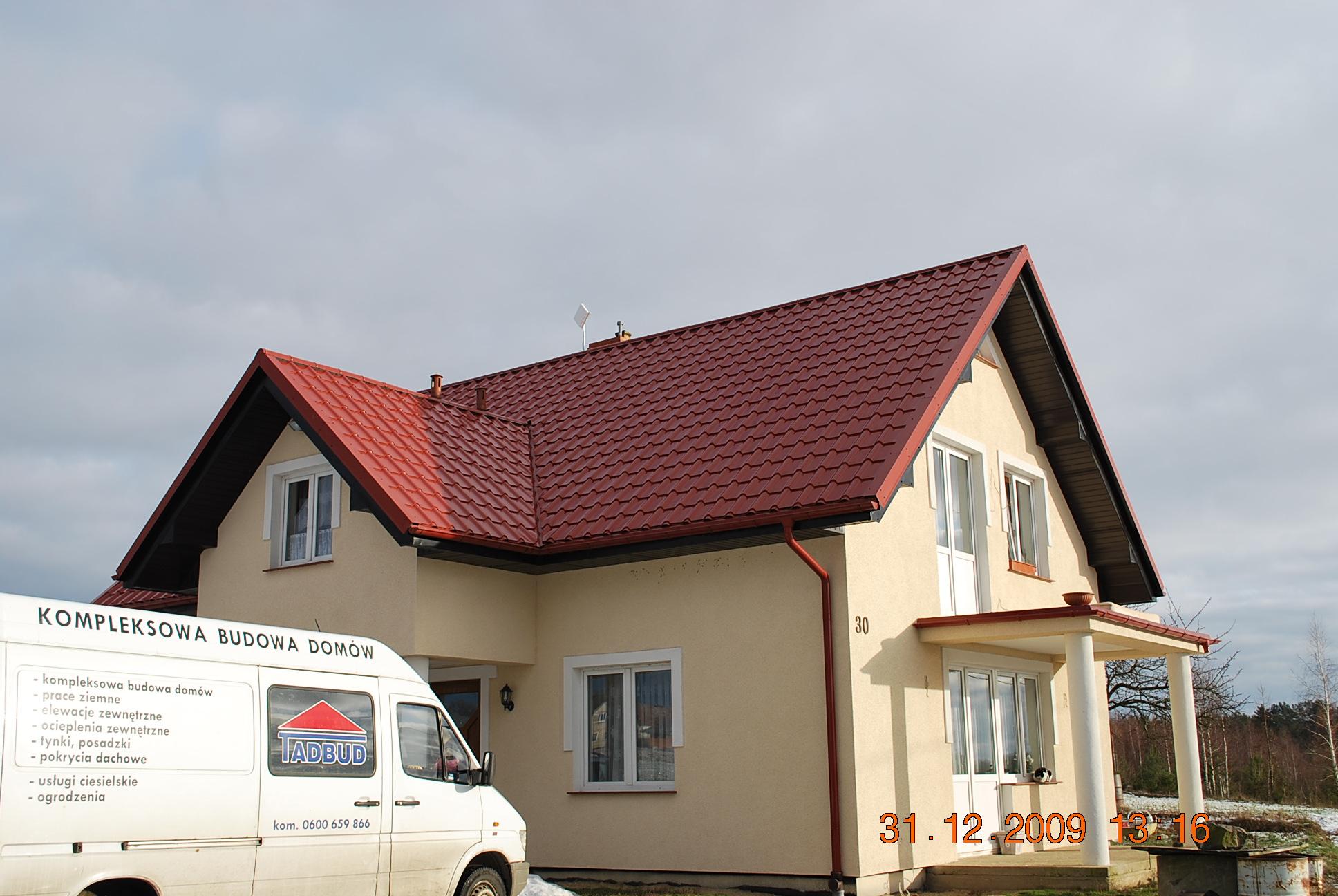 Dom jednorodzinny z nowym, czerwonym dachem z dachówki, widoczny z boku, z samochodem firmy budowlanej z logo IADBUD zaparkowanym przed budynkiem. Na dachu widoczna antena.